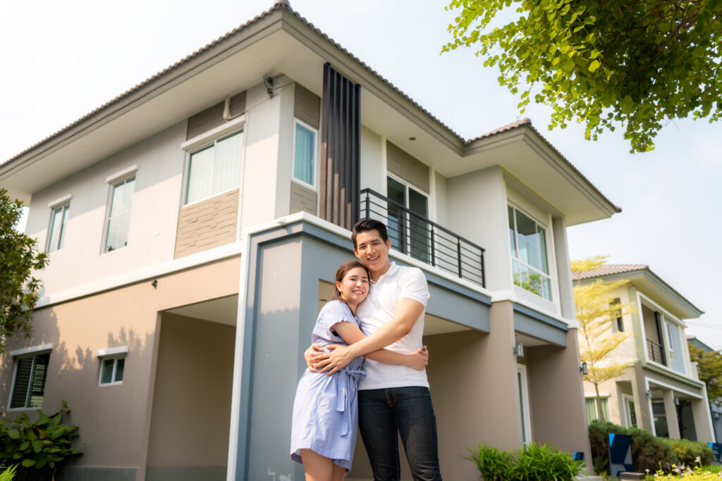 Portrait of Asian young couple standing and hugging together looking happy in front of their new house to start new life. Family, age, home, real estate and people concept.