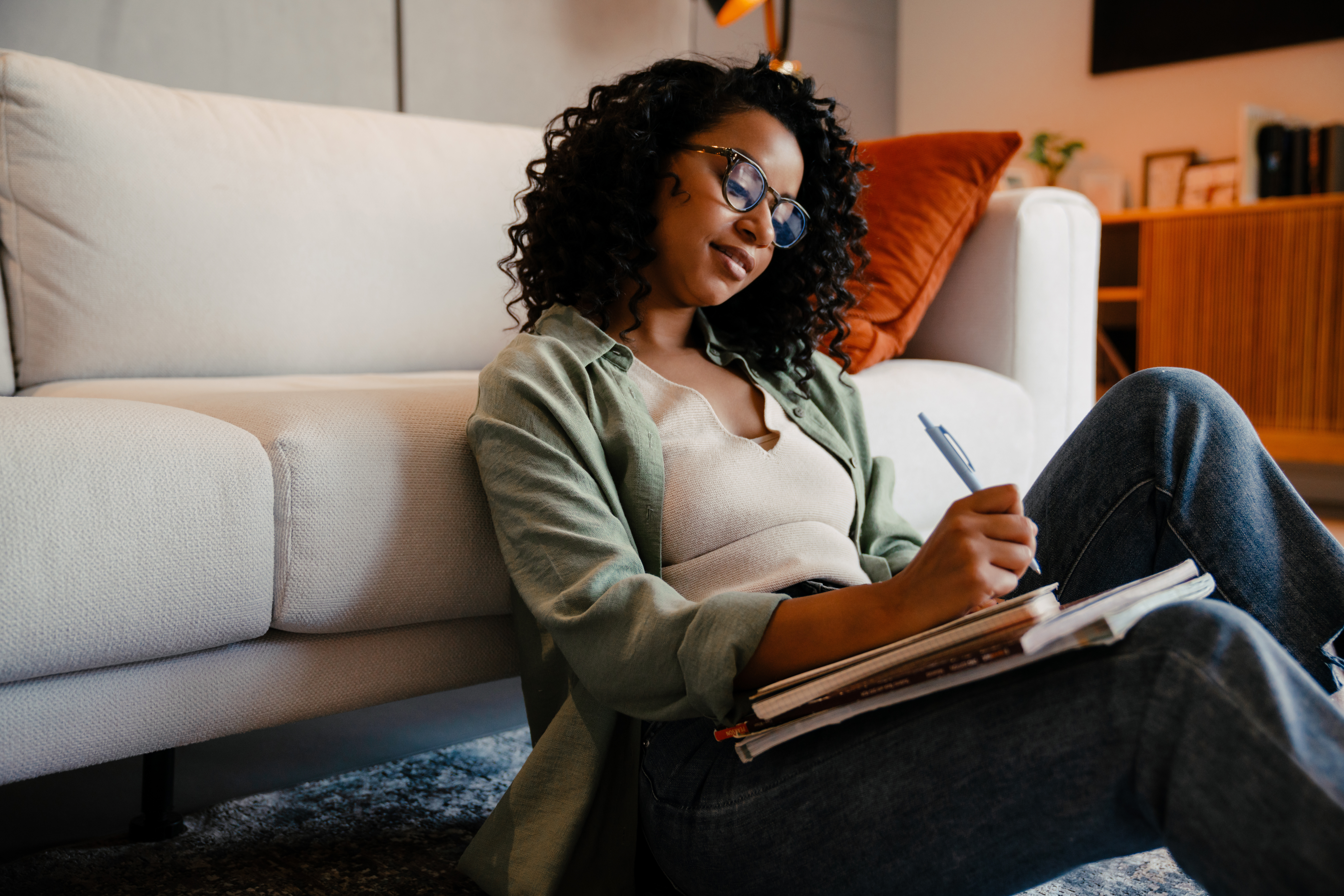 Young african woman in eyeglasses writing down notes while sitting on the floor at home